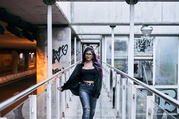 stock image latina girl with long hair glasses brunette fur jacket ripped jeans pierced lip walking on the glass walkway - Urban concept