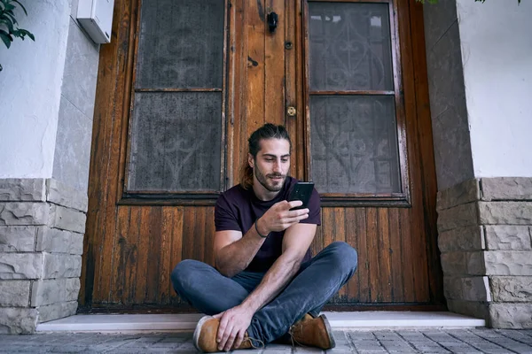 Caucasian young man with long hair and beard touching smartphone screen of him smiling sitting by the wooden door of the house - Urban style