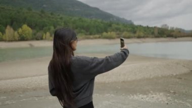 Gözlüklü güzel bir kız kendini cep telefonuyla göl kenarında filme çekiyor.