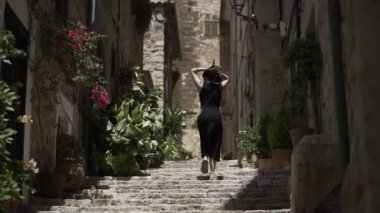 Genç bir İspanyol kadın güneşli bir günde taştan merdivenlerden çıkarken saçlarını topluyor. - Paradisiac İspanyol mekanları