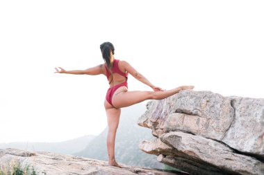 Beyaz, olgun bir kadın spor yapıyor, sol bacağı yerde, sağ bacağı ise büyük kayanın üstünde.