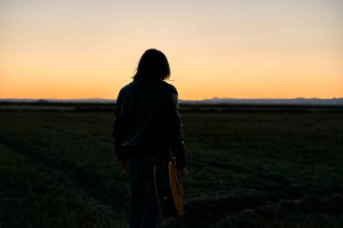 Kafkasyalı, uzun saçlı, büyük yeşil bir tarlada elinde İspanyol gitarıyla günbatımında maço bir yaşam tarzıyla duran bir çocuk.