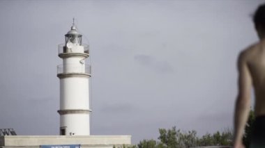 Bulutlu bir akşamda yavaş çekimde deniz fenerine doğru yürüyen genç üstsüz bir adam. - Paradisiac İspanyol mekanları