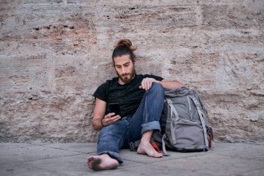 Kafkas gezgin çocuk sakin ve yalın ayak görünüyor ve akıllı telefonuna dokunuyor büyük sırt çantasına yaslanıyor kirli ayaklarıyla duvarın yanında