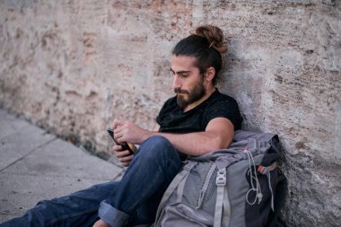 Uzun saçlı genç beyaz adam yerde, taş bir duvara yaslanmış büyük bir sırt çantasının yanında oturup akıllı telefonunun ekranına bakarak sakal topladı.