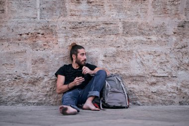 Sakallı, beyaz, kirli ayaklı, çıplak ayaklı, taş bir duvara yaslanmış, elinde akıllı telefonuyla sırt çantasıyla gezen genç bir adam.