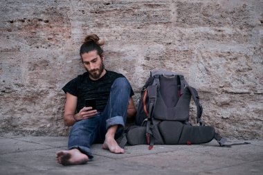 Yalınayak yorgun Kafkas çocuk akıllı telefonuna bakıyor ve dokunuyor duvara yaslanıyor sırt çantasının yanında kirli ayaklarıyla sırt çantasıyla