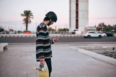 Çizgili kazaklı ve kafasında koruyucu kaskla elinde kaykayla akıllı telefonuna bakan beyaz bir genç.