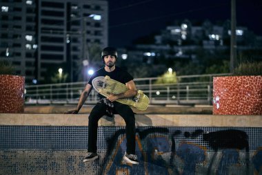 Genç beyaz kaykaycı, kafasına kask takmış göğsünde kaykayıyla şehir dışındaki duvarda sakin ve rahat bir şekilde oturuyor.