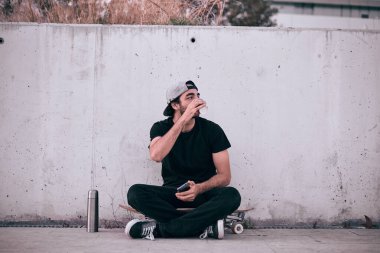 Gri şapkalı, siyah tişörtlü, kaykaylı beyaz bir adam elinde akıllı telefonuyla bir bardaktan kahve içiyor.