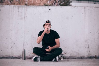 Beyaz şapkalı, siyah tişörtlü, kaykaylı beyaz bir adam termosun yanında oturmuş duvarında akıllı telefon olan bir bardaktan kahve içiyor.