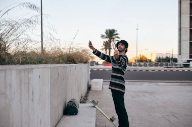 Çizgili süveter giymiş, burnuna piercing takmış, elinde akıllı telefonuyla kameraya bakan, şehirde kaykay yapan beyaz bir adam.