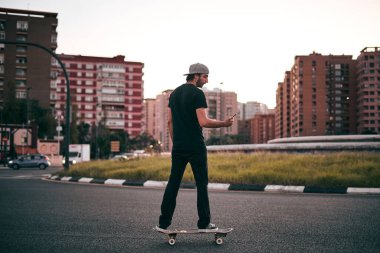 Siyah tişörtlü ve şapkalı genç beyaz adam kaykayıyla asfaltta sürerken akıllı telefonunun ekranına bakıyor.