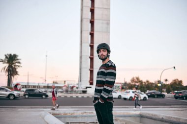 Çizgili kazaklı ve kafasında koruyucu miğfer olan genç beyaz bir adam gürültülü bir şehirde elleri ceplerinde sokakta dikiliyor.