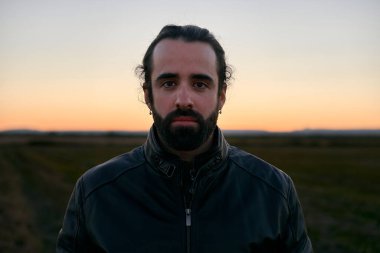 Saçları toplayan ve sakalları düzgün beyaz bir adam gün batımında düğmeli bir kürk ceketle kameraya ciddi ciddi bakıyor.