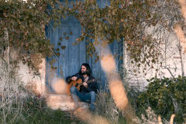 Kürk ceketli genç beyaz bir adam bir evin kapısında oturuyor ve gözleri kapalı tutkuyla gitar çalıyor.