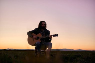 Beyaz, uzun saçlı ve sakallı genç bir adam, tahta bir taburede oturuyor ve büyük yeşil bir tarlada gitar çalıyor.