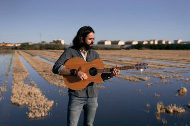 Sakallı ve küpeli genç beyaz adam gün batımında su altında kalmış bir tarlada İspanyol gitarı çalıyor.
