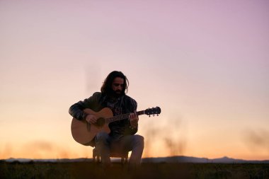 Kafkasyalı genç adam, uzun saçlı, alanın ortasında bir sandalyede oturuyor günbatımında gitar çalıyor, maço yaşam tarzı.