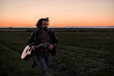 Beyaz, uzun saçlı, sakallı ve ceketli bir genç gün batımında elinde gitarıyla çayırlarda koşuyor.