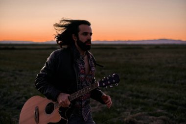 Gün batımında elinde gitarıyla çayırlarda koşuşturan uzun saçlı ve kürklü beyaz bir delikanlı.