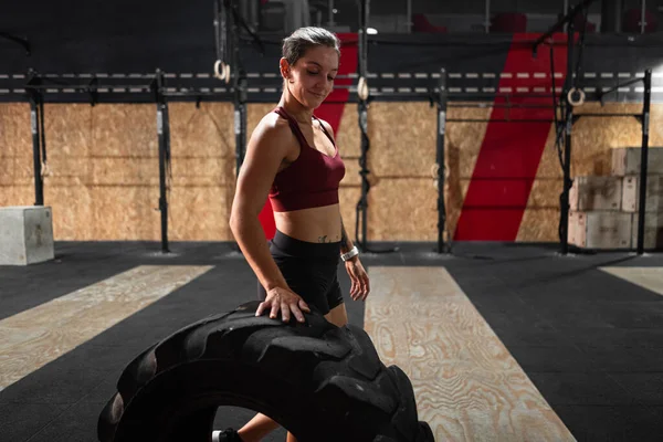 stock image caucasian girl athlete standing smiling rolling a big crossfit tractor wheel in a big gym - Crossfit concept