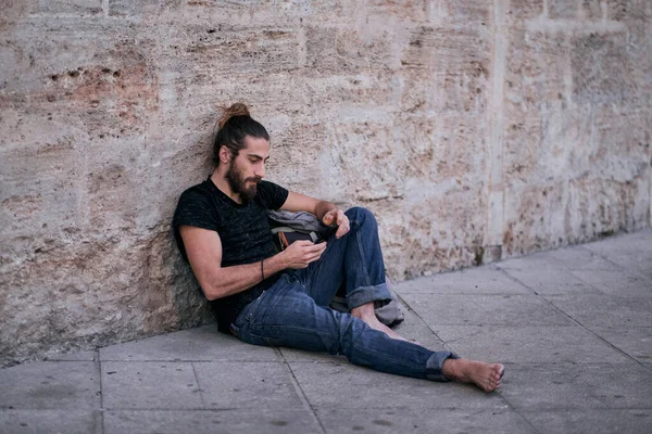 Caucasian young man with long hair barefoot calm and relaxed sitting on the floor next to a large backpack leaning against a stone wall looking at the screen of his smartphone - Backpacker travel