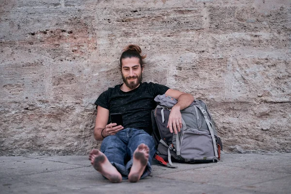 caucasian guy with long hair legs stretched out barefoot looking at camera with his smartphone in hand sitting leaning on his big backpack on the floor by the wall with dirty feet - Backpacker travel