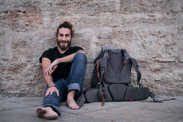 Beyaz sakallı, uzun saçlı, çıplak ayaklı, mutlu ve memnun bir adam sırt çantasının yanında kirli ayaklarıyla duvara yaslanmış, sırt çantasıyla geziyor.