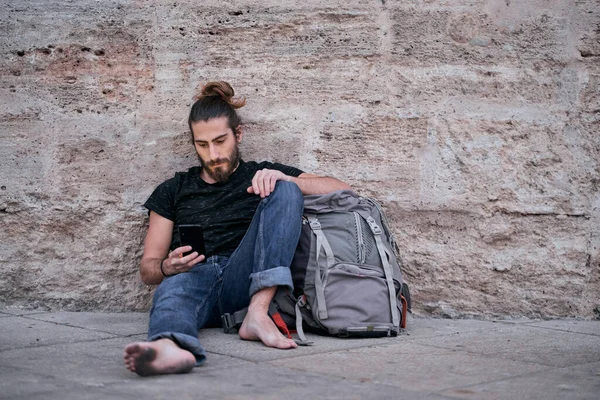 barefoot tired traveler caucasian guy looking and touching his smartphone sitting leaning on his big backpack on the floor by the wall with dirty feet - Backpacker travel concept