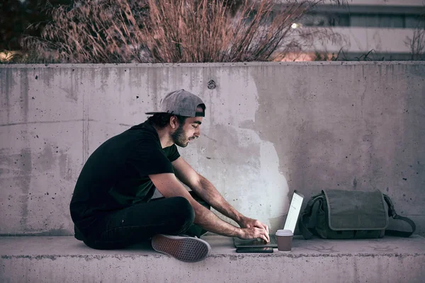 caucasian guy in gray cap black t-shirt sitting on the concrete floor with his legs crossed manipulating his laptop at sunset in the city - Lfestyle concept