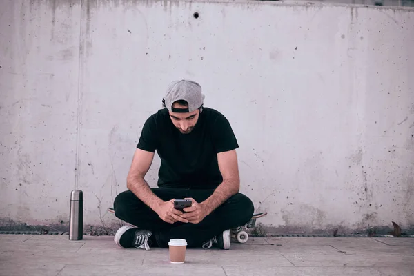 caucasian guy in a gray cap and black t-shirt sitting on his skateboard manipulating his smartphone next to a thermos and a glass of coffee - Lfestyle concept