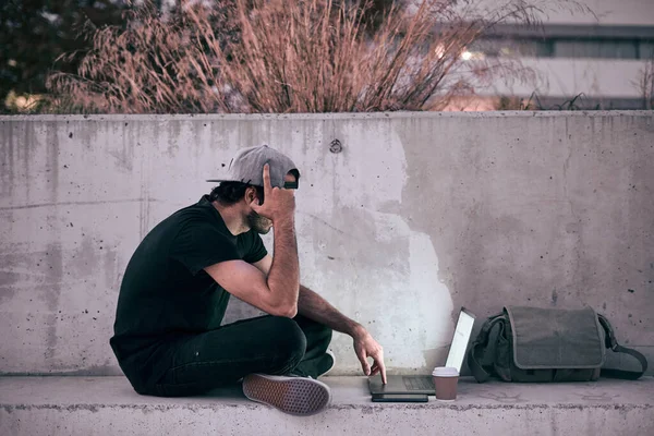 caucasian guy in gray cap black t-shirt sitting on the concrete floor with his legs crossed looking at his laptop screen next to the smartphone drinking coffee - Lfestyle concept