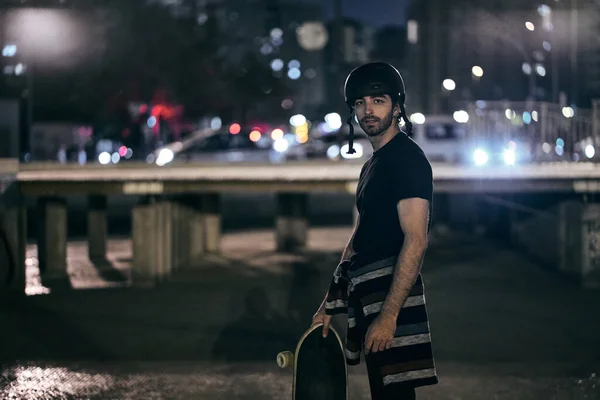 caucasian skateboarder guy in a striped jumper tied at the waist holding a skateboard with his hand in the city night standing looking at camera - Lfestyle concept