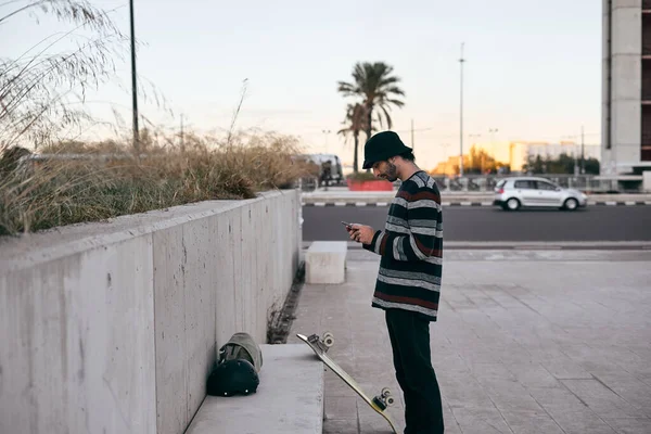 줄무늬 스웨터를 피어싱하는 코카서스 남자가 스케이트보드 스타일 도시에서 스마트폰 화면을 — 스톡 사진