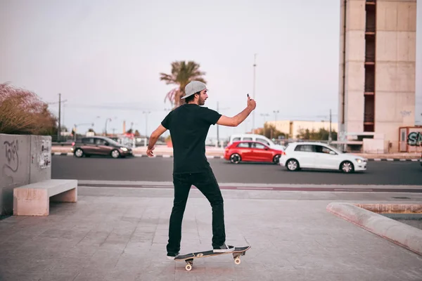 黒のTシャツを着た白人の若者と頭の上に帽子彼のスケートボードのバランスをとる彼のスマートフォンで自撮りを取る ライフスタイルの概念 — ストック写真