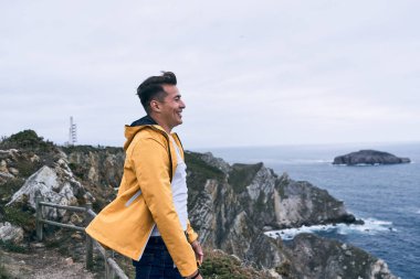 Beyaz tişörtlü ve sarı ceketli latin genç adam sakin ve rahat bir şekilde deniz kenarında durup temiz hava alıyor, Cabo de Penas, İspanya - İspanya manzaraları