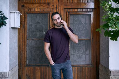 Beyaz sakallı genç adam ahşap kapı ve posta kutusunun yanında akıllı telefonuyla konuşuyor.