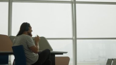 Havaalanı terminalinde kulaklık takan genç beyaz bir adam arka planda uçan bir uçak - dijital göçebe konsepti