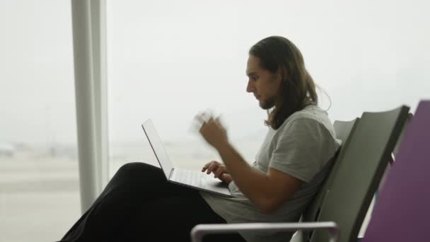 年轻的长发男子用他的笔记本电脑坐在机场 中镜头 数字游牧民族的概念 — 图库视频影像