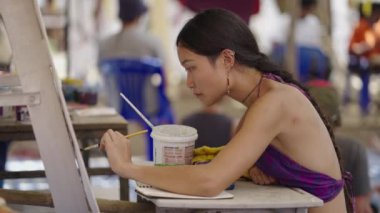 Kalabalık bir yerde Asyalı bir kadın tuvale resim çiziyor. Hippi bir sanatçı.