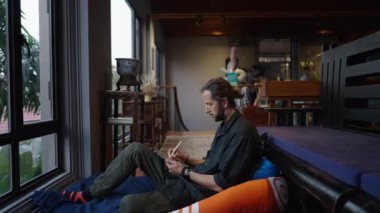 Sakallı bohem bir adam tabletini etnik olarak dekore edilmiş bir odada çalıyor.