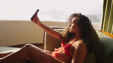 Güzel esmer kadın güneşli pencerenin yanında selfie çekiyor.