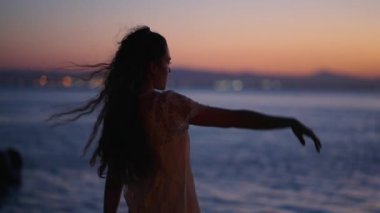 Sırtı dönük güzel bir kız gün batımında kollarını kaldırıyor - 4K Yatay video