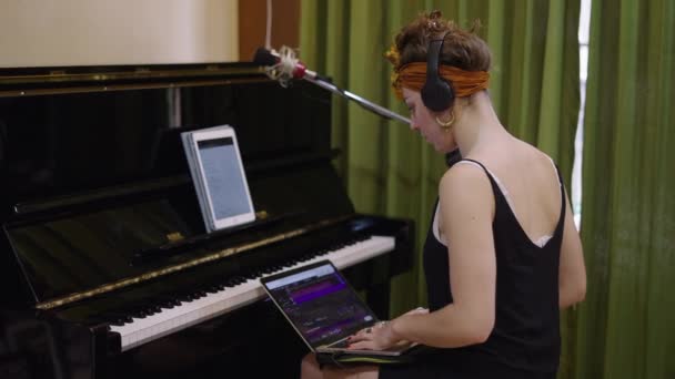 女孩坐在钢琴前 用她的耳机从笔记本电脑上听音乐 4K水平视频 — 图库视频影像