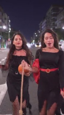 Cadı kostümlü iki güzel Kolombiyalı kız, biri elinde balkabağı, Valencia, İspanya 'da gece sokakta yürürken eğleniyor. - Tam Vuruş - Dikey 1080
