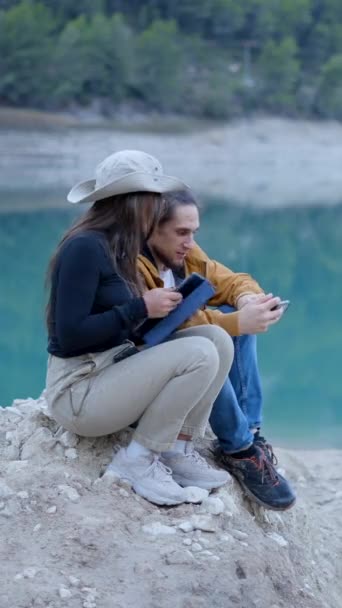 ハッピーカップルは水とチャットで岩の上に座って タブレットと電話を使用します 垂直ビデオ — ストック動画