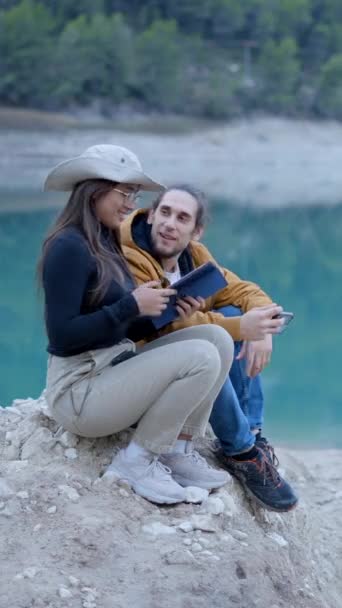 若いカップルはまだ湖で岩の上に座って話し キスしてタブレットを使用します 垂直ビデオ — ストック動画