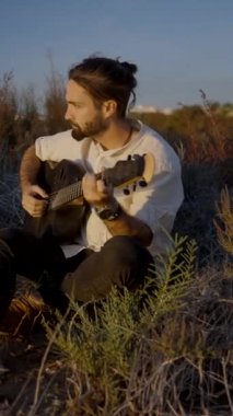 Sakallı İspanyol adam gün batımında akustik gitar çalıyor düşünceli bir yüz ifadesi var.