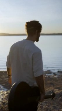 Güneş batarken Torrevieja Pembe Gölü 'nde yürüyen gitarlı genç bir adam, Alicante, Kırsal İspanya - arkası görünümü, dikey FullHD videosu